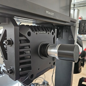 Op de afbeelding: Een zwarte race stuurbevestiging met een zilveren metalen as en een zwart schuimgevoerd uiteinde. De bevestiging is gemarkeerd met "Fanatec" en is bevestigd aan een zwart metalen frame.