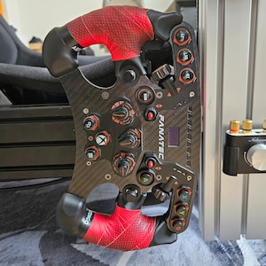 Op de afbeelding: Een zwart race stuur met rode accenten en een carbon fiber afwerking. Het stuur heeft knoppen, peddels en een display. De tekst "FANATEC" is zichtbaar op het stuur.