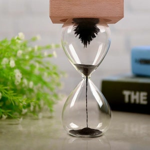 May include: A clear glass hourglass with black sand, a wooden top, and a magnetic pendulum inside. The hourglass is on a white surface.