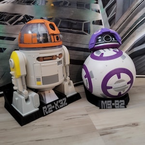 May include: Two Star Wars droid figures, R2-X3Z and MB-82, sit on black bases. R2-X3Z is white, orange, and yellow. MB-82 is white and purple. The bases have the droid names printed on them.