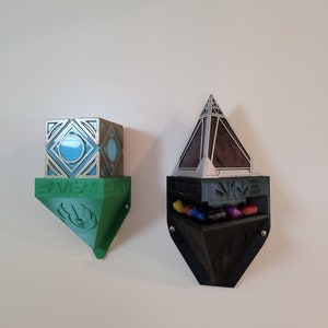 May include: Two 3D printed wall mounted display shelves. The left shelf is green and shaped like a triangle with a lightsaber symbol. The right shelf is black and shaped like a pyramid with a small compartment for holding items. The compartment is filled with colourful items.