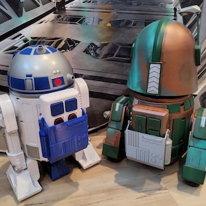 May include: Two toy robots, one white and blue, the other green and brown. The white robot has a silver head with blue and red accents. The green robot has a helmet-like head with a brown and green color scheme. Both have rectangular compartments.