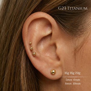 May include: A close-up of an ear with multiple gold-colored ball stud earrings. The image includes text that reads "G23 TITANIUM", "16g 18g 20g", and measurements of 5mm, 6mm, 8mm, and 10mm.