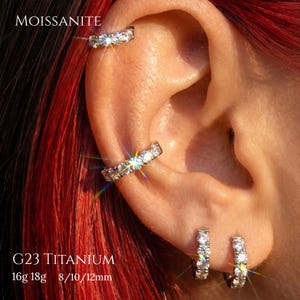 May include: A set of silver-toned G23 titanium hoop earrings and a cartilage cuff, each featuring a row of sparkling round moissanite stones. The jewelry is displayed on an ear, with text reading MOISSANITE, G23 TITANIUM, 16g 18g, and 8/10/12mm.