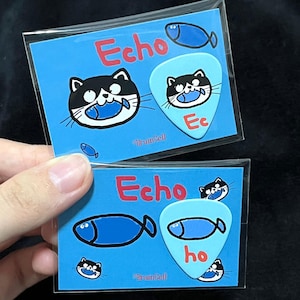 Peut inclure: Deux autocollants bleus pour médiators de guitare avec un chat de dessin animé tenant un poisson, le mot "Echo" en rouge et le texte "Braincell". Un médiator a la forme d'un poisson et l'autre est un médiator de guitare.