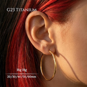 Op de afbeelding: Een goudkleurige oorring gemaakt van G23 titanium. De oorring is cirkelvormig met een gladde, gepolijste afwerking. Tekst op de afbeelding specificeert G23 TITANIUM, 16g 18g en 20/30/40/50/60mm, wat de beschikbare diktes en diameters voor dit sieraad aangeeft.