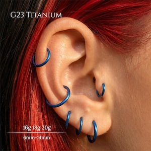 Puede incluir: Un conjunto de seis aros circulares y lisos de titanio azul G23 usados en varios piercings de oreja. El texto G23 TITANIUM, 16g 18g 20g y 6mm-14mm es visible en la imagen, indicando el material, los calibres y el rango de diámetros.