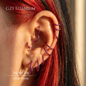 Puede incluir: Una colección de aros de titanio G23 de color morado usados en múltiples perforaciones de oreja. El texto G23 TITANIUM, 16g 18g 20g y 6mm-14mm es visible. Estos anillos circulares son delgados, lisos y metálicos, con un acabado morado vibrante.