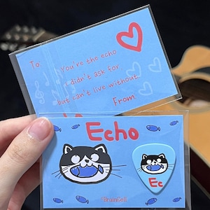 Peut inclure: Un ensemble de cartes et de médiators bleu clair. Le médiator présente un chat noir et blanc tenant un poisson. La carte dit : "Tu es l'écho que je n'ai pas demandé, mais sans lequel je ne peux pas vivre." Le médiator porte les lettres "Ec".