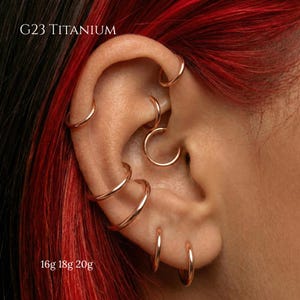 Peut inclure: Gros plan sur une oreille ornée de plusieurs boucles d'oreilles créoles en or rose. L'image présente différentes tailles et emplacements des boucles d'oreilles, avec le texte "G23 TITANIUM" et "16g 18g 20g" visibles. L'oreille est posée sur un fond de cheveux roux.