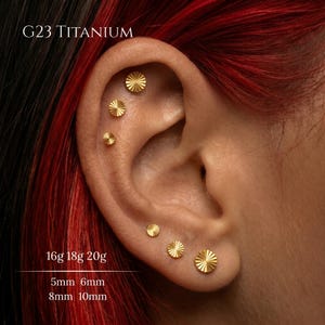 Puede incluir: En una oreja se muestran varios pendientes de titanio dorados de diferentes tamaños. Los pendientes tienen un diseño de rayos de sol. El texto de la imagen dice "G23 TITANIUM" y los tamaños son 5 mm, 6 mm, 8 mm y 10 mm.