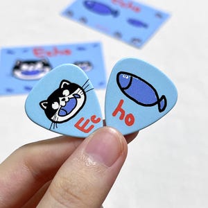 Peut inclure: Deux médiators de guitare bleu clair avec des illustrations de dessins animés. L'un représente un chat noir et blanc avec le mot "Echo" en rouge. L'autre a un poisson bleu et le mot "ho" en rouge. Cartes bleues supplémentaires avec des poissons et des chats en arrière-plan.