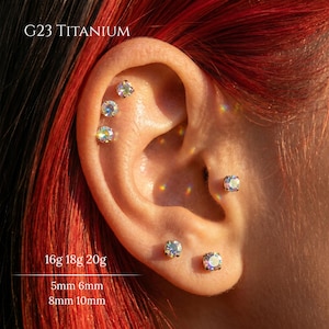 May include: Close-up of an ear adorned with multiple iridescent titanium earrings. The text "G23 TITANIUM" is visible at the top. Below, the text indicates sizes: 16g 18g 20g and 5mm 6mm 8mm 10mm. The earrings are round and square.