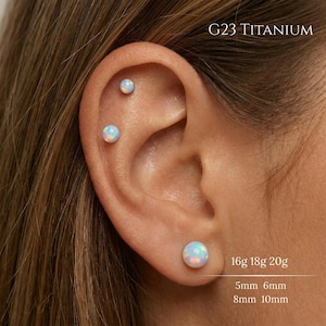 Puede incluir: Primer plano de una oreja con tres pendientes de ópalo. Los pendientes son redondos y tienen un aspecto nacarado e iridiscente. El texto de la imagen dice "G23 TITANIUM" y "16g 18g 20g" con medidas de 5 mm, 6 mm, 8 mm y 10 mm.