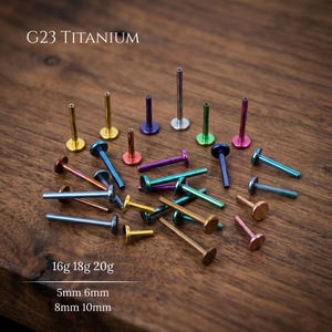 Op de afbeelding: Een collectie G23 titanium labret-studs in diverse metallic kleuren, waaronder goud, blauw, paars, groen en roségoud. De tekst G23 TITANIUM, 16g 18g 20g en 5mm 6mm 8mm 10mm is zichtbaar. Deze lichaamsieraden zijn voorzien van platte achterkantjes.