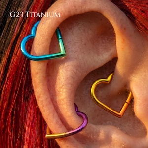 Può includere: Tre orecchini a cerchio a forma di cuore in titanio G23, di colore blu, giallo e viola, indossati in diversi piercing alla cartilagine. I cerchi in metallo presentano una finitura liscia e lucida con un design a segmento incernierato.