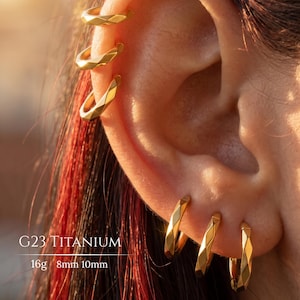 May include: A set of gold-coloured hoop earrings with a faceted geometric texture. The jewellery is labelled as G23 Titanium with a 16g gauge and 8mm or 10mm diameter options. The earrings are shown worn in multiple piercings along the ear cartilage and lobe.