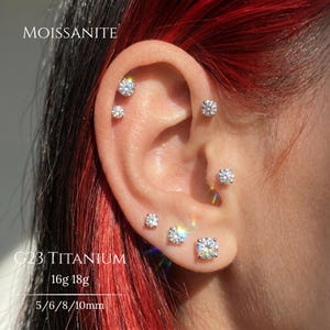 May include: A set of six round, clear moissanite stud earrings in silver-toned G23 titanium settings, worn in multiple ear piercings. The text MOISSANITE, G23 TITANIUM, 16g 18g, and 5/6/8/10mm is visible on the image, indicating material and sizes.