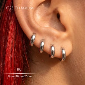 Può includere: Quattro orecchini a cerchio in titanio G23 color argento, disponibili nelle misure 8mm, 10mm e 12mm con uno spessore di 16g. Gli anelli in metallo liscio e lucido sono disposti in fila sul lobo, riflettendo la luce con una finitura metallica.