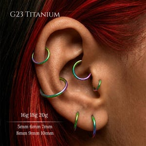 May include: An ear with multiple piercings, each adorned with iridescent hoop earrings. The image displays several piercings with different hoop sizes. The text "G23 TITANIUM" is visible, along with size details: 16g 18g 20g and 5mm 6mm 7mm 8mm 9mm 10mm.