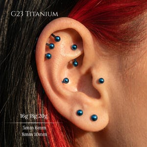 May include: A collection of G23 titanium body jewelry pieces featuring metallic blue spherical ends. The text G23 TITANIUM, 16g 18g 20g, and 5mm 6mm 8mm 10mm is visible. These small circular studs and curved bars are placed in various ear piercings.
