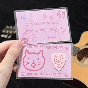 Puede incluir: Juego de púas y tarjetas de guitarra rosas con diseño de gato. La tarjeta dice "A little practice goes a long way" y tiene una ilustración de gato. La púa tiene una ilustración de gato y la marca "BrainCell".