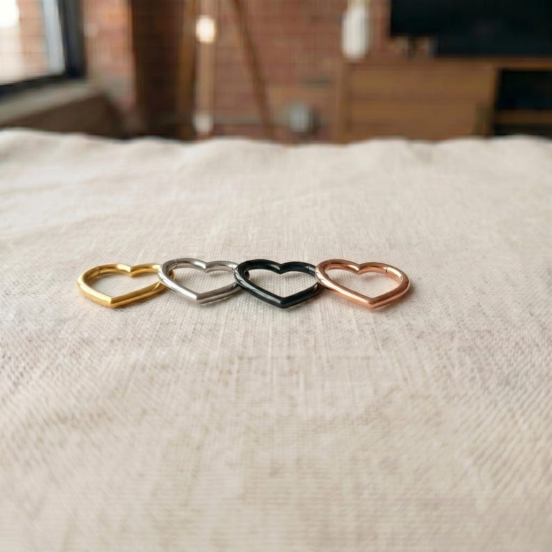 Pode incluir: Quatro argolas em forma de cora&ccedil;&atilde;o dispostas numa fila horizontal. O conjunto inclui uma argola em tom dourado, uma em tom prateado, uma preta e uma em tom de ouro rosa. Cada argola possui um mecanismo de fecho com dobradi&ccedil;a.