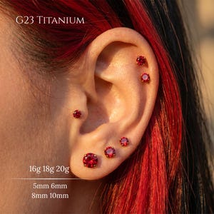 Könnte beinhalten: Ein Ohr mit mehreren Piercings, jedes mit einem kleinen, runden, roten Edelstein in goldfarbenem Metall. Der Text "G23 TITANIUM" steht oben. Darunter steht "16g 18g 20g" und "5mm 6mm 8mm 10mm".