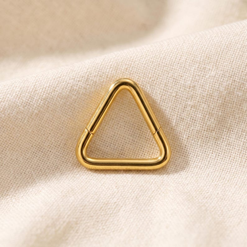 Peut inclure: Une boucle d'oreille cr&eacute;ole en m&eacute;tal dor&eacute; poli en forme de triangle. La pi&egrave;ce pr&eacute;sente une surface lisse et continue avec une couture visible l&agrave; o&ugrave; le segment articul&eacute; rencontre le cadre principal, cr&eacute;ant une esth&eacute;tique g&eacute;om&eacute;trique &eacute;pur&eacute;e.