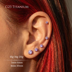 May include: Close-up of an ear with multiple purple gemstone stud earrings. The text "G23 TITANIUM" is at the top. The image also shows the sizes: 16g 18g 20g, 5mm 6mm, and 8mm 10mm.