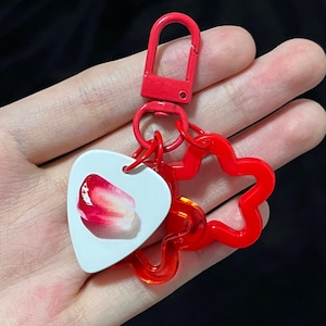 May include: A red keychain with a lobster clasp, a flower-shaped charm, and a white guitar pick-shaped charm. The guitar pick charm has a close-up image of a strawberry slice. The keychain is held in a hand.