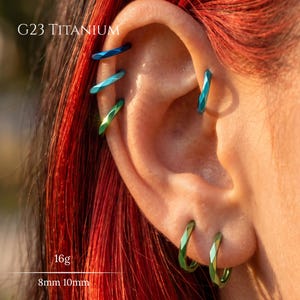 May include: G23 titanium hoop earrings in 16g with 8mm and 10mm diameters. These metallic hoops feature a faceted, angular surface texture and transition in color from deep blue to teal and green, worn in multiple ear piercings.