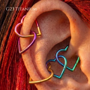 May include: G23 TITANIUM heart-shaped hoop earrings in a vibrant gradient of purple, pink, gold, and teal. These metallic hinged rings feature a smooth finish and are designed for ear piercings, showcasing a colorful spectrum across the metal surface.