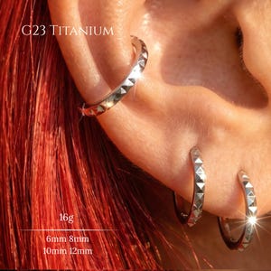 Puede incluir: Tres pendientes de aro de titanio G23 en color plateado con un patrón texturizado de tachuelas piramidales. El texto G23 TITANIUM, 16g y 6mm 8mm 10mm 12mm es visible. Los aros tienen un acabado metálico y se llevan en la oreja, brillando.