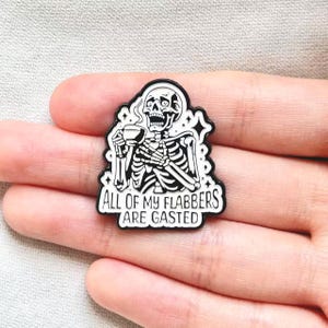 Op de afbeelding: Zwart-witte emaille pin met een skelet dat een kopje koffie vasthoudt. Het skelet heeft een verbaasde uitdrukking en de tekst "ALL OF MY FLABBERS ARE GASTED" staat eronder.