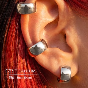 Peut inclure: Trois créoles en titane G23 argenté de tailles variées portées sur une oreille. Le texte G23 TITANIUM 18g 8mm 10mm est visible. Ces boucles d'oreilles circulaires lisses et réfléchissantes présentent un design à bande épaisse et une finition métallique brillante.