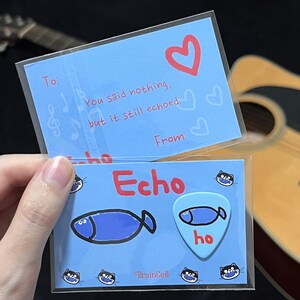 Peut inclure: Une carte bleu clair avec le texte "You said nothing, but it still echoed." et un cœur rouge. En dessous, une carte avec le mot "Echo" et un dessin de poisson bleu. Un médiator avec un dessin de poisson et le mot "ho".