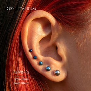 May include: Five G23 titanium ball stud earrings arranged in a row along the earlobe. The studs feature a metallic blue finish, except for the largest one which is silver. Text on the image reads G23 TITANIUM, 16g 18g 20g, and 5mm 6mm 8mm 10mm.