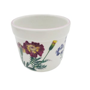 May include: A white ceramic flower pot with a light pink rim. The pot is decorated with colorful floral illustrations, including a large maroon and yellow flower, green leaves, and blue flowers. Ideal for planting flowers or herbs.