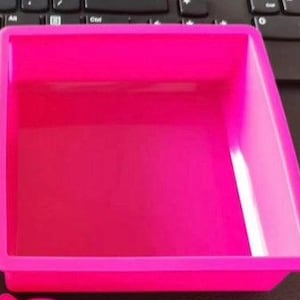 Op de afbeelding: Een felroze, rechthoekige siliconen mal met licht afgeronde hoeken. De mal is leeg en staat op een donkere ondergrond, waarschijnlijk een bureau of tafel. De algemene vorm is een eenvoudige, open bak.