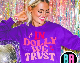 Könnte beinhalten: Lilafarbener Pullover mit dem Text "In Dolly We Trust" in rosa und roten Buchstaben. Der Pullover ist cropped und hat eine lockere Passform. Das Model trägt ein weißes Stirnband und ein silbernes Armband.