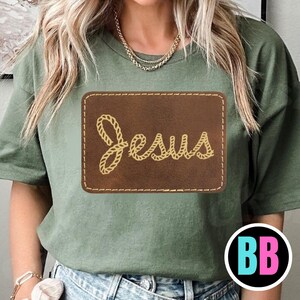 May include: Olive green t-shirt with a brown rectangular patch featuring the word "Jesus" in a rope-like font. The patch is stitched with a contrasting thread. The shirt has short sleeves and a crew neck.