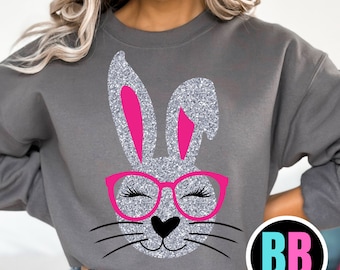 Può includere: Una felpa grigia con un coniglio glitterato che indossa occhiali rosa. Il coniglio ha un naso rosa e baffi. La felpa ha un logo "BB" in basso a destra.