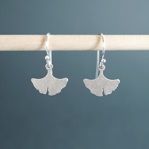 May include: A pair of silver ginkgo leaf earrings with a textured finish. The earrings are hanging from a wooden dowel.
