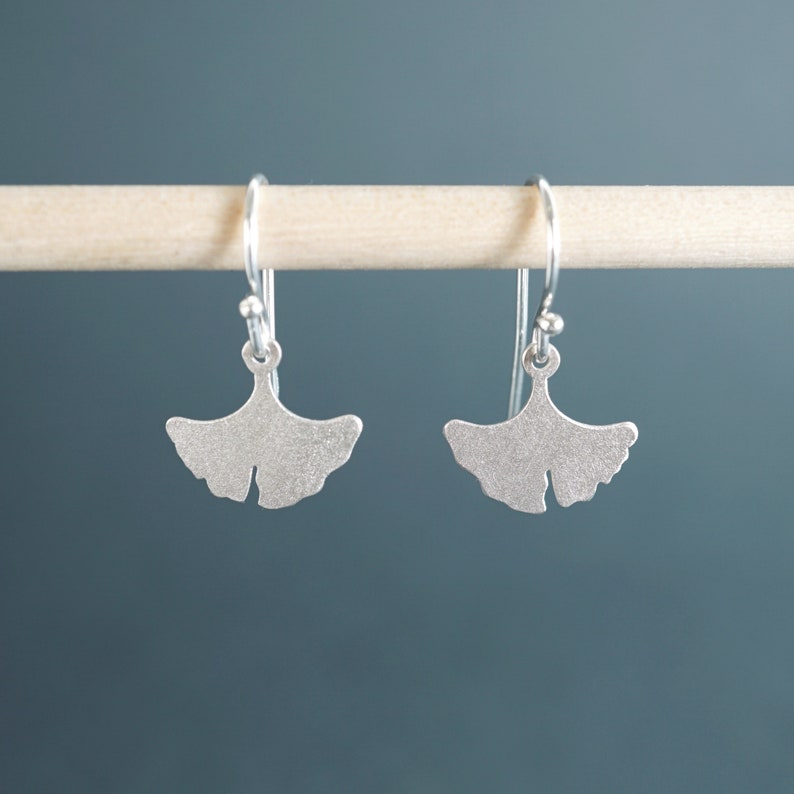 May include: A pair of silver ginkgo leaf earrings with a textured finish. The earrings are hanging from a wooden dowel.