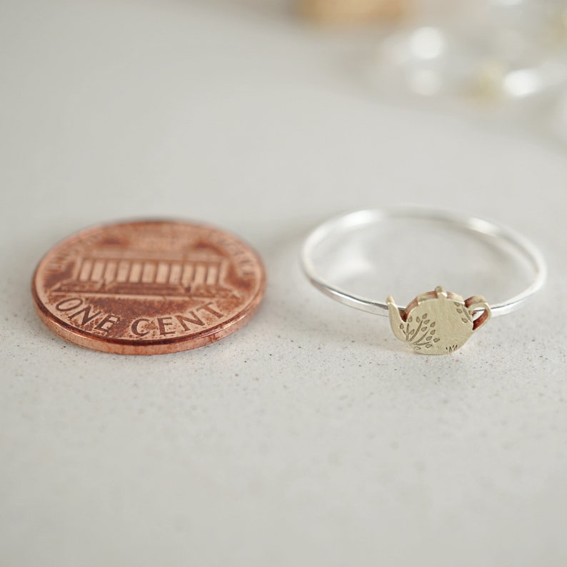 K&ouml;nnte beinhalten: Ein silberner Ring mit einem goldenen Teekannen-Anh&auml;nger. Die Teekanne hat ein florales Design und ist etwa so gro&szlig; wie ein deutscher Cent.