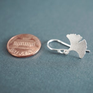 May include: A silver ginkgo leaf earring with a hook closure. The earring is shown next to a US penny for size comparison.