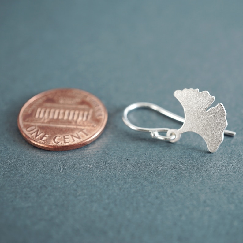 May include: A silver ginkgo leaf earring with a hook closure. The earring is shown next to a US penny for size comparison.