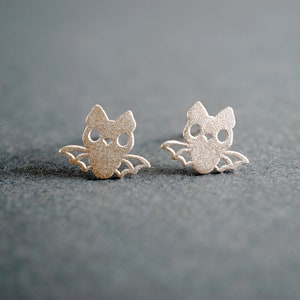 Peut inclure: Une paire de boucles d'oreilles en argent en forme de chauves-souris. Les chauves-souris ont de petits yeux ronds et des ailes.