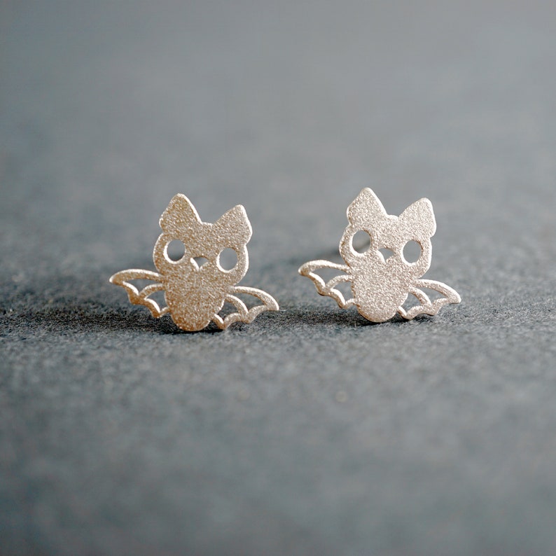 Peut inclure: Une paire de boucles d'oreilles en argent en forme de chauves-souris. Les chauves-souris ont de petits yeux ronds et des ailes.